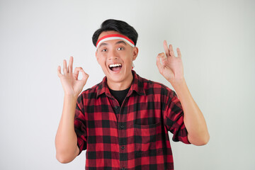 Excited young asian man wearing Indonesian flag headband, smiling expression, hand raised with okay gesture, isolated over white background. Concept for Indonesian Independence Day.