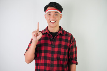 Smiling young asian man wearing Indonesian flag headband and pointing fingers on copy space upside, isolated over white background. Concept for Indonesian Independence Day.