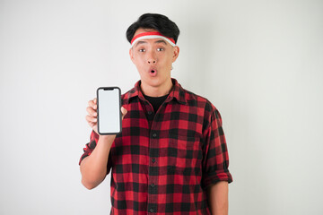 Smiling young asian man wearing Indonesian Flag headband is showing vertical blank smartphone screen for advertising, isolated over white background. Indonesian Independence Day.