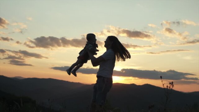Silhouette of happy young mother spins pretty baby son on background mountains during sunset. Playful mom and toddler boy playing on field at summertime. Family having fun together. Slow-motion