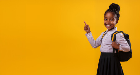 Smiling black school girl pointing her forefinger at an empty space for promotional text, yellow background. Schoolgirl with a big smile. Back to school sales concept