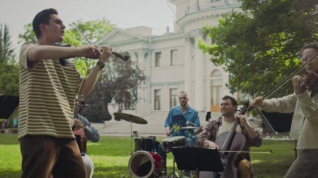 Group of young classical musicians playing cellos, violins and drums giving performance while busking in park