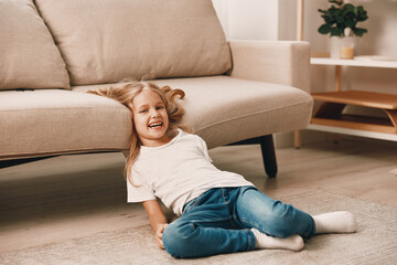 Sad little girl sitting on the floor next to a couch with her head in her hands in a cozy living...