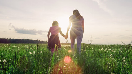 Mother and her little daughter running holding hands across the beautiful green grassland at sunset.