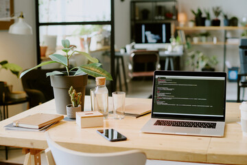 No people shot of modern laptop with code on screen, plants in pots, smartphone and glass bottle of water on desk in eco-friendly office