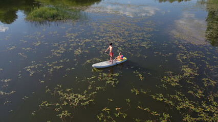 Two children enjoying summer holiday and sunny day, paddling stand up board on a calm lake water,...
