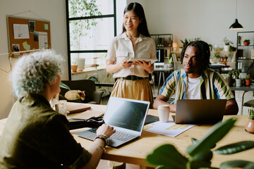 Cheerful young Asian manager having conversation with African American and Caucasian programmers at work in modern office