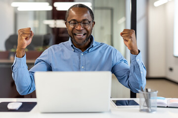 African American office worker celebrating success at workplace inside office. Mature professional man wearing glasses smiling and raising fists in joy and accomplishment.