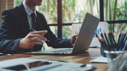 A businessman at a modern wooden desk focusing intently on his laptop, surrounded by charts and statistical reports in a bright, green-filled office.