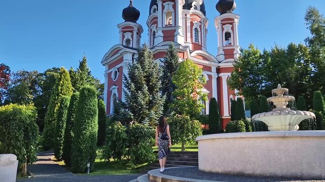Young woman walks in the gardens of Curchi Monastery with outdoor view to the water fountain. Christian Orthodox church with a beautiful green yard located in Orhei, Moldova