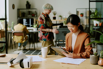 Young Asian manager sitting at desk using digital tablet at work in modern office, her colleagues printing documents and coding in background