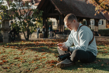 Fototapeta premium Professional Photographer Taking Picture Of Beautiful Autumn Park. Man Professional Photographer Sit With Camera And With Smartphone In Autumn Park. Retouched, Vibrant Colors, Brownish Tones.