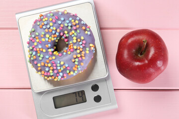 Kitchen scale with donut and apple on pink wooden table, top view