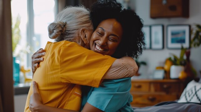 A young woman in blue scrubs hugs an elderly woman in a yellow shirt. They are both smiling warmly and appear to be very happy.