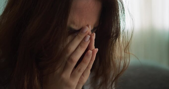 Close up a brunette girl covers herself with her hands and cries at home near the sofa. The face of a brunette girl who cries after a sad conversation