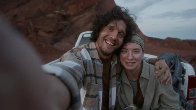 Medium shot of young Caucasian couple sitting together, hugging in pickup truck on stopover during offgrid journey, enjoying tea, taking selfie on smartphone, smiling, waving, looking at camera