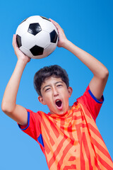 A kid raises a soccer ball upwards with his hands celebrating a goal. Isolated on blue background....