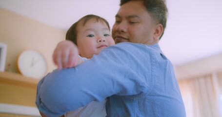 Obraz premium A Asian father holding his little toddler child on shoulder in a bright living room, capturing a moment of family bonding and happiness.