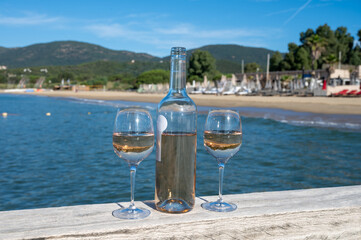 Bottle and glass of cold rose wine from Provence and wooden yacht boota pier on white sandy beach Plage de Pampelonne near Saint-Tropez, summer vacation in France