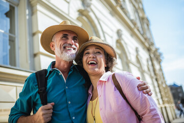 Old married couple exploring the city. Hugging each other. They are wearing hats and backpacks.