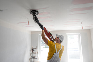 The worker sanding a plasterboard ceiling. He is using special drywall sander. A red plasterboard...