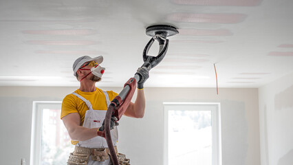 The worker sanding a plasterboard ceiling. He is using special drywall sander. A red plasterboard improve the fire resistance of ceiling structures.