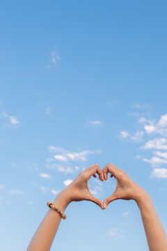 A woman raises her hand above her head to make a heart symbol on the background of the bright morning sky because the heart symbol signifies love, friendship and kindness to one another.