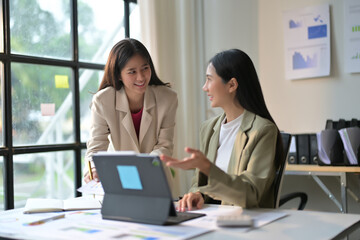 Obraz premium Two Businesswomen Collaborating in Modern Office with Laptop and Documents, Discussing Work Strategies