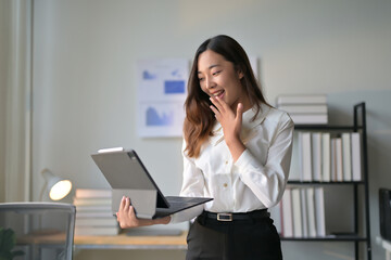 Young Professional Woman Excitedly Looking at Laptop in Modern Office Setting with Bookshelves and Charts in Background