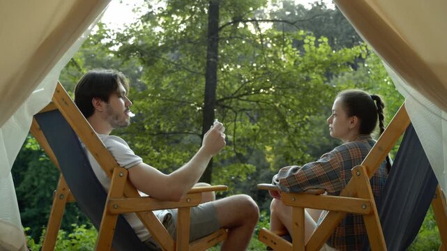 Young couple is seen vaping in nature, sitting in chairs in front of their tent. Medium shot. Slow motion.
