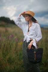 Fashionable confident woman wearing trendy summer outfit with wide brim hat, white linen shirt, green skirt, holding black wicker  straw bag, posing outdoor.  Fashion portrait