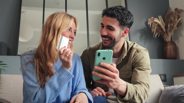 A young couple ordering food online using a delivery app on a mobile phone. Man and woman are sitting on the couch in their house, paying online with a credit card and a cellphone for a trip.