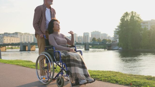 Senior woman in wheelchair walking with caregiver old man on road in park. Elderly family couple man husband supporting embracing woman wife in chair for people with disability outdoor. Rehabilitation