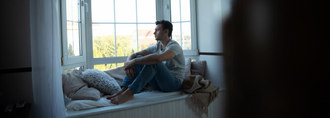 Fototapeta premium Depressed, sad, frustrated, crying, scared, instability mental and bipolar young man sitting on the windowsill at home. Headache because overworked and stress