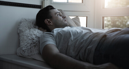 Side view photo of handsome young laying down man on the windowsill on the pillow and looking out of the window and thinking about something. Relaxing at home after hard work