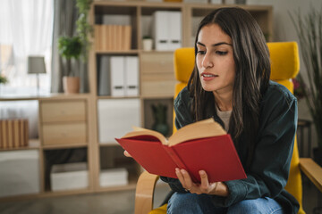 Adult young woman sit on a chair and read a book in the living room