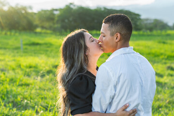 young latin couple, kissing while strolling through a field of green grass. sunset in the background