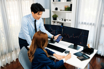 Smiling businessman showing dynamic stock market data on mobile phone to woman writing to memo for...