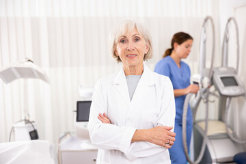 Smiling old woman doctor posing against background of cosmetology devices in aesthetic clinic