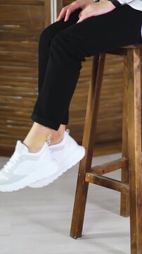 Woman sits on a high stool waving her feet in white sneakers. Model jumps off the tabouret and walks away in comfortable footwear. Vertical video