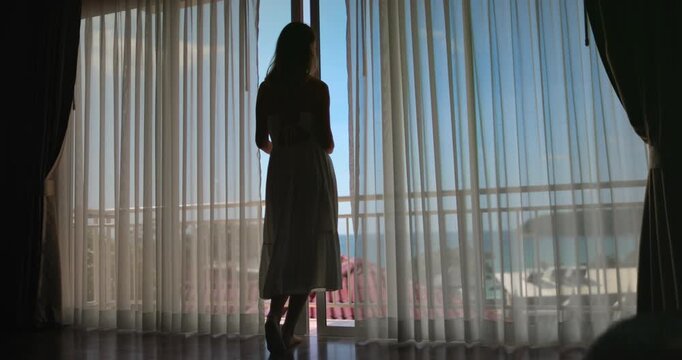 Young woman is silhouetted against a bright window overlooking a tropical beach as she opens the curtains in her hotel room. Enjoy the beauty of travel and vacation with this evocative image