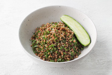 White bowl with boiled buckwheat and kale on a gray background, served with fresh cucumber. Healthy homemade vegan food