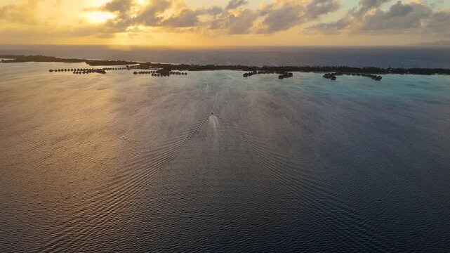 A small commuter boat travels over south pacific ocean water near the tropical island of Bora Bora to overwater bungalow resorts during the early morning sunrise. Aerial drone view. 