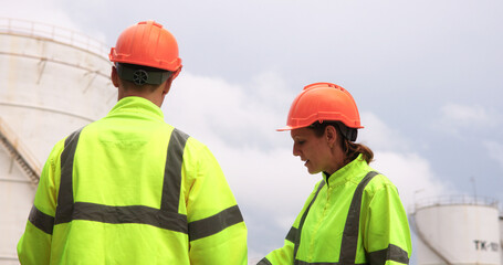 Male and female energy engineers collaborate with a laptop, blueprint, and digital tablet at the oil storage tanks in chemical and refinery  plant