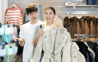 Young woman buyer in store has removed hanger with fur coat from window and lay heads together with...