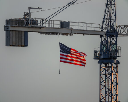 United States flag gracefully flying, back lit by setting sun and hanging from crane on a construction site.