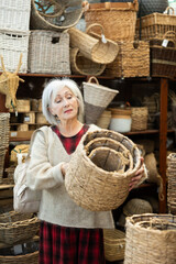 Senior woman buyer in store of homemade self-made goods made of osier shoot, wicker decorative items considers large and small rattan baskets for storing items