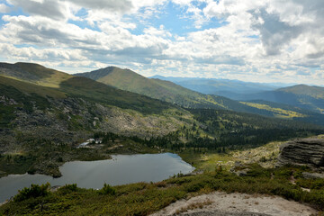 A view from the top to a picturesque lake lying in an intermountain basin surrounded by peaks overgrown with a rare coniferous forest under a cloudy summer sky.