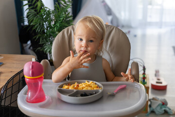 a blonde baby eats with a spoon while sitting at a children's dining table