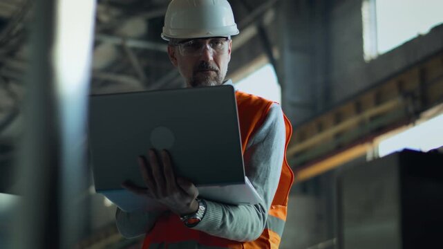 Professional heavy industry male specialist in protective uniform, hardhat and goggles stand at manufacturing facility or factory. Mature engineer or technician works using laptop computer. Close Up.